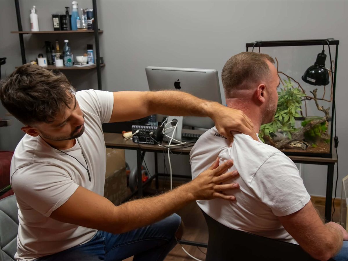 Masér Marek pri masáži ramien klienta, ktorý sedí pri pracovnom stole s počítačom